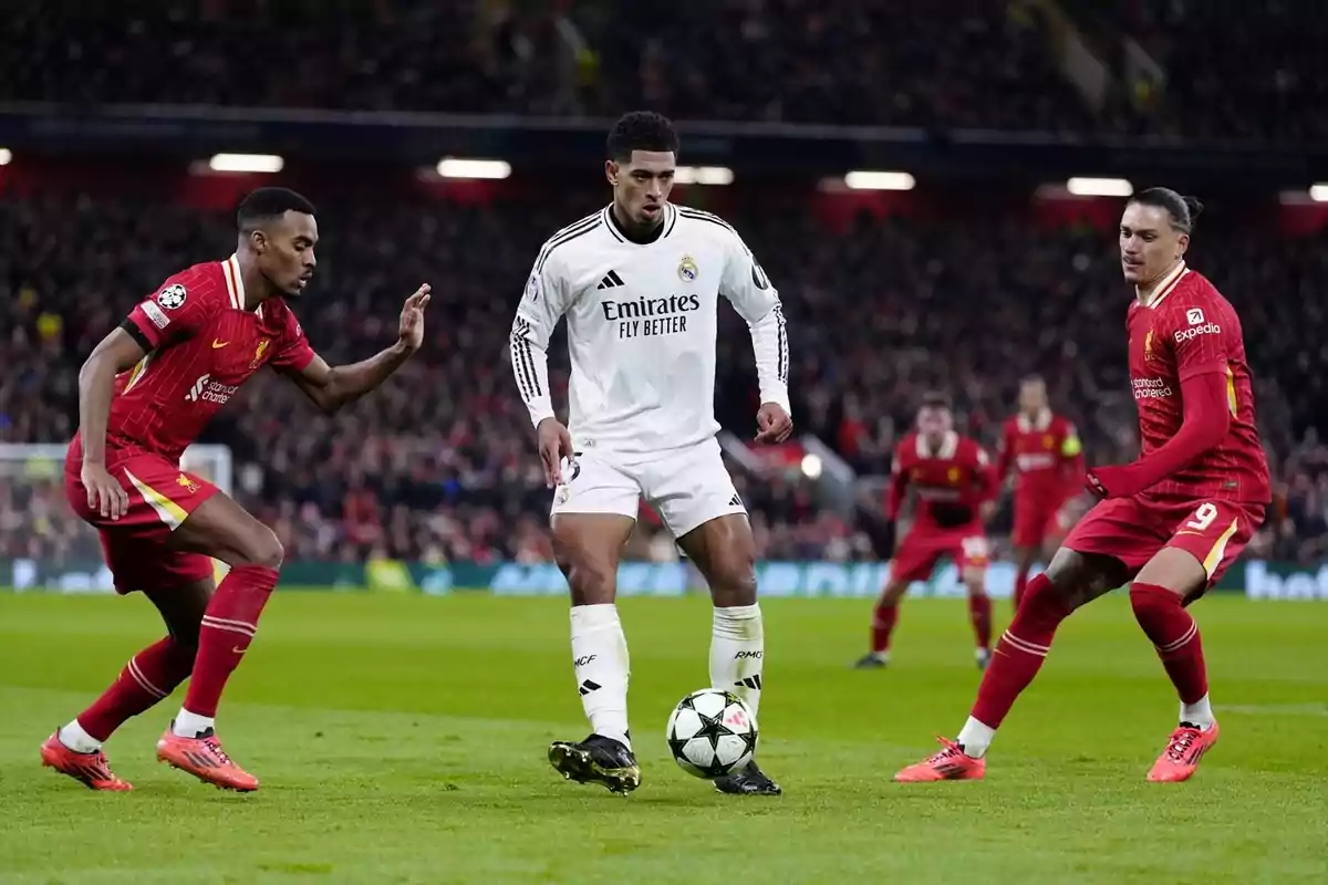 Un jugador del Reial Madrid controla la pilota mentre dos futbolistes del Liverpool intenten prendre-li la pilota durant un partit de futbol en un estadi ple d’espectadors. Un jugador del Reial Madrid controla la pilota mentre dos futbolistes del Liverpool intenten prendre-li la pilota durant un partit de futbol en un estadi ple d’espectadors.