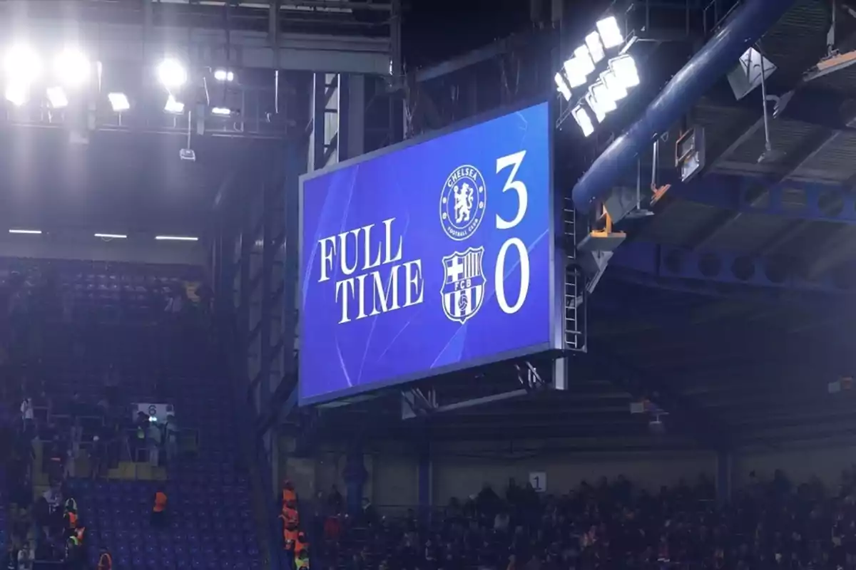 Marcador final en una pantalla gigante mostrando la victoria de Chelsea 3 a 0 sobre Barcelona en un estadio de fútbol con público en las gradas.