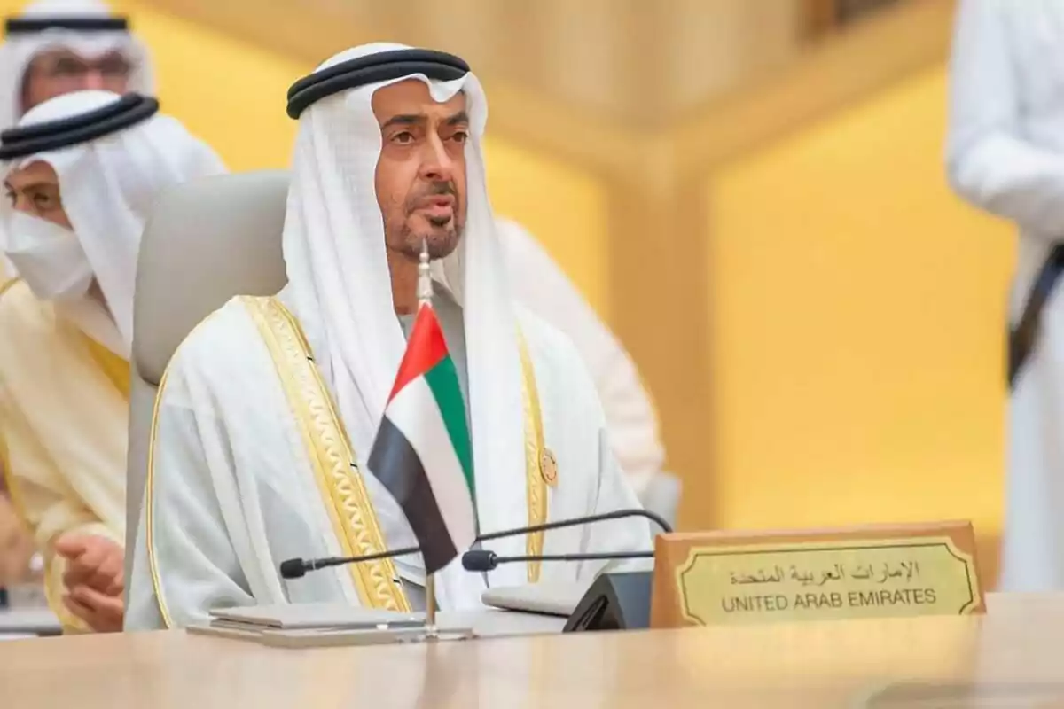Mohamed bin Zayed, con vestimenta tradicional árabe, sentado en una conferencia con una bandera de Emiratos Árabes Unidos frente a él. Mohamed bin Zayed, con vestimenta tradicional árabe, sentado en una conferencia con una bandera de Emiratos Árabes Unidos frente a él.