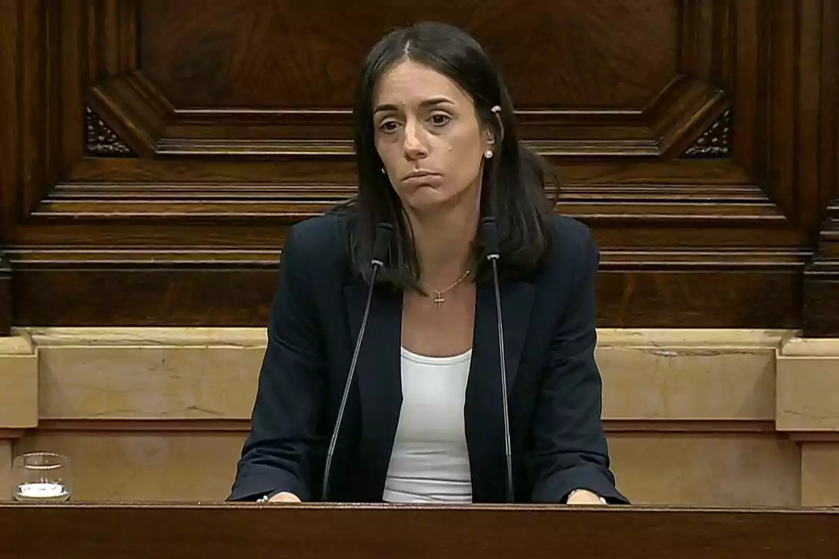 Mujer de cabello oscuro y expresión seria hablando en un podio con dos micrófonos en un entorno institucional de madera