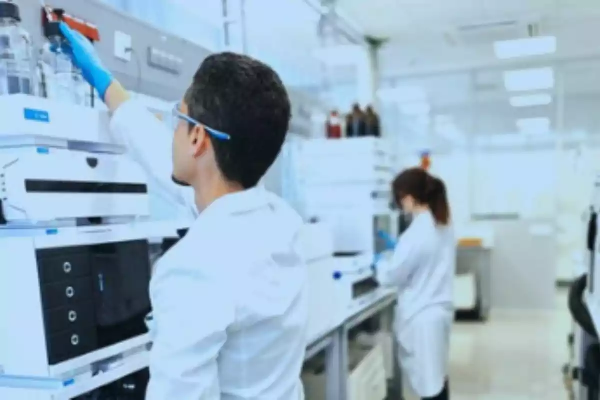 Dos personas con bata de laboratorio y guantes trabajan en un laboratorio moderno rodeados de equipos científicos. Dos personas con bata de laboratorio y guantes trabajan en un laboratorio moderno rodeados de equipos científicos.