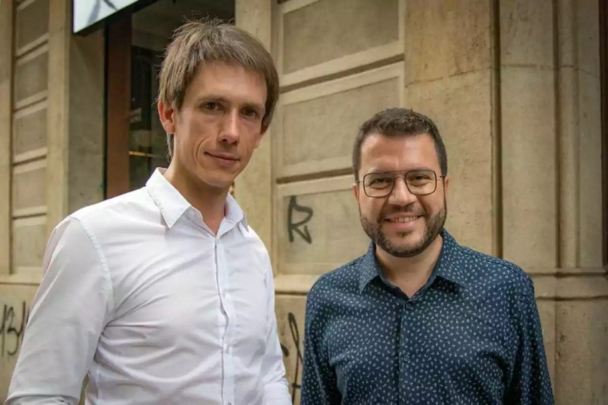 Dos hombres posan juntos y sonríen frente a una pared de piedra en una calle urbana