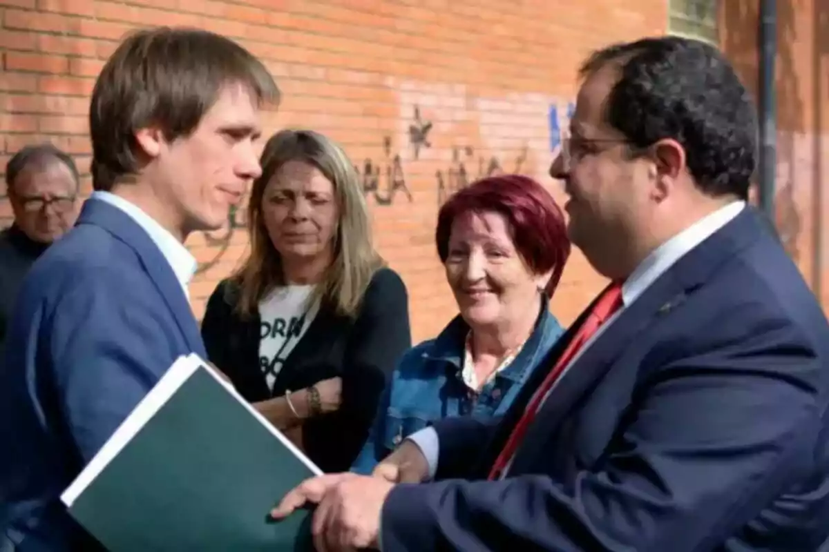 Dos hombres de traje conversan y se dan la mano mientras dos mujeres observan y sonríen en un entorno urbano con una pared de ladrillo al fondo