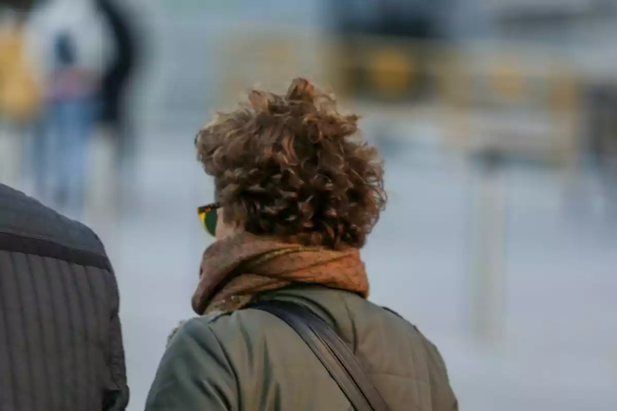 Persona de espaldas con cabello rizado usando bufanda marrón y chaqueta verde en un entorno urbano desenfocado. Persona de espaldas con cabello rizado usando bufanda marrón y chaqueta verde en un entorno urbano desenfocado.