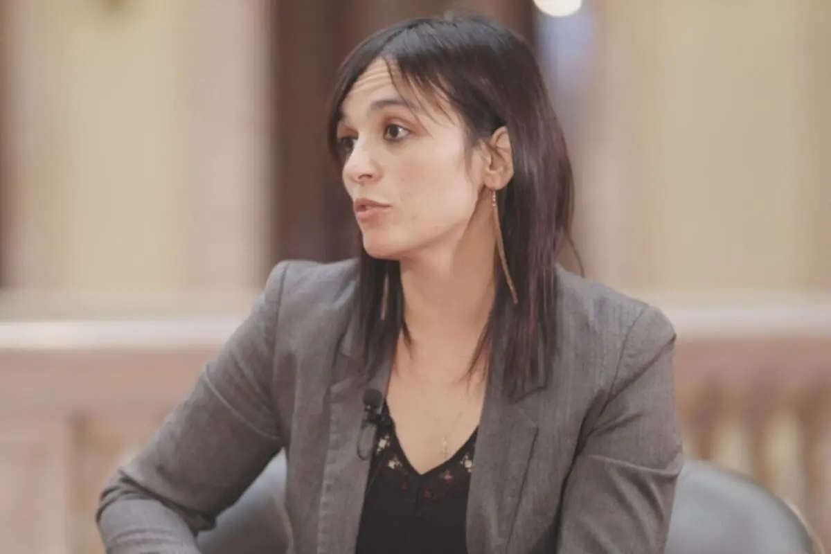 Mujer de cabello oscuro y lacio usando saco gris sentada en un ambiente interior mientras habla