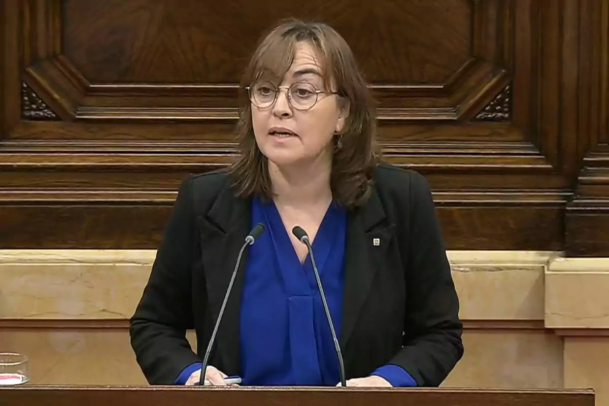 Dona amb ulleres i jaqueta negra parlant en un faristol amb fons de fusta Dona amb ulleres i jaqueta negra parlant en un faristol amb fons de fusta