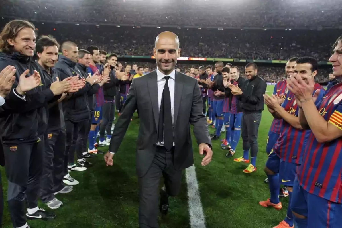 A soccer coach walks smiling past a row of players who applaud him in a stadium full of spectators.