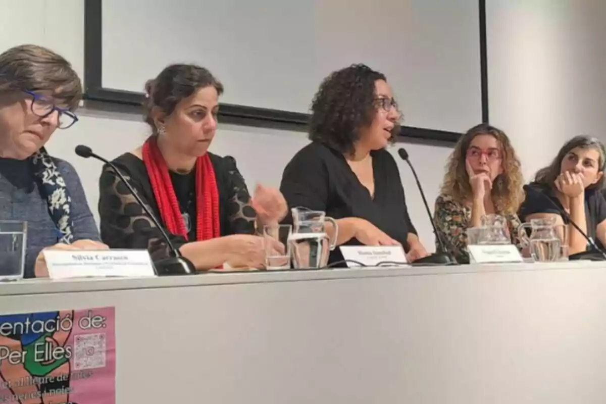 Cinco mujeres sentadas en una mesa durante una conferencia o panel, con micrófonos y jarras de agua frente a ellas, en un ambiente interior y profesional.