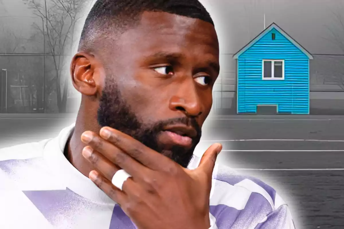 Hombre con barba y camiseta blanca con detalles morados tocándose la barbilla mientras al fondo se ve una casa azul