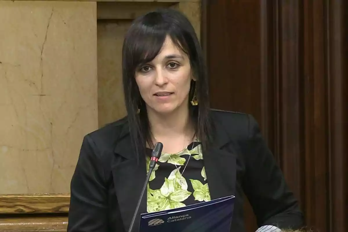 Mujer de cabello oscuro y liso hablando frente a un micrófono en un entorno formal con una carpeta azul en las manos