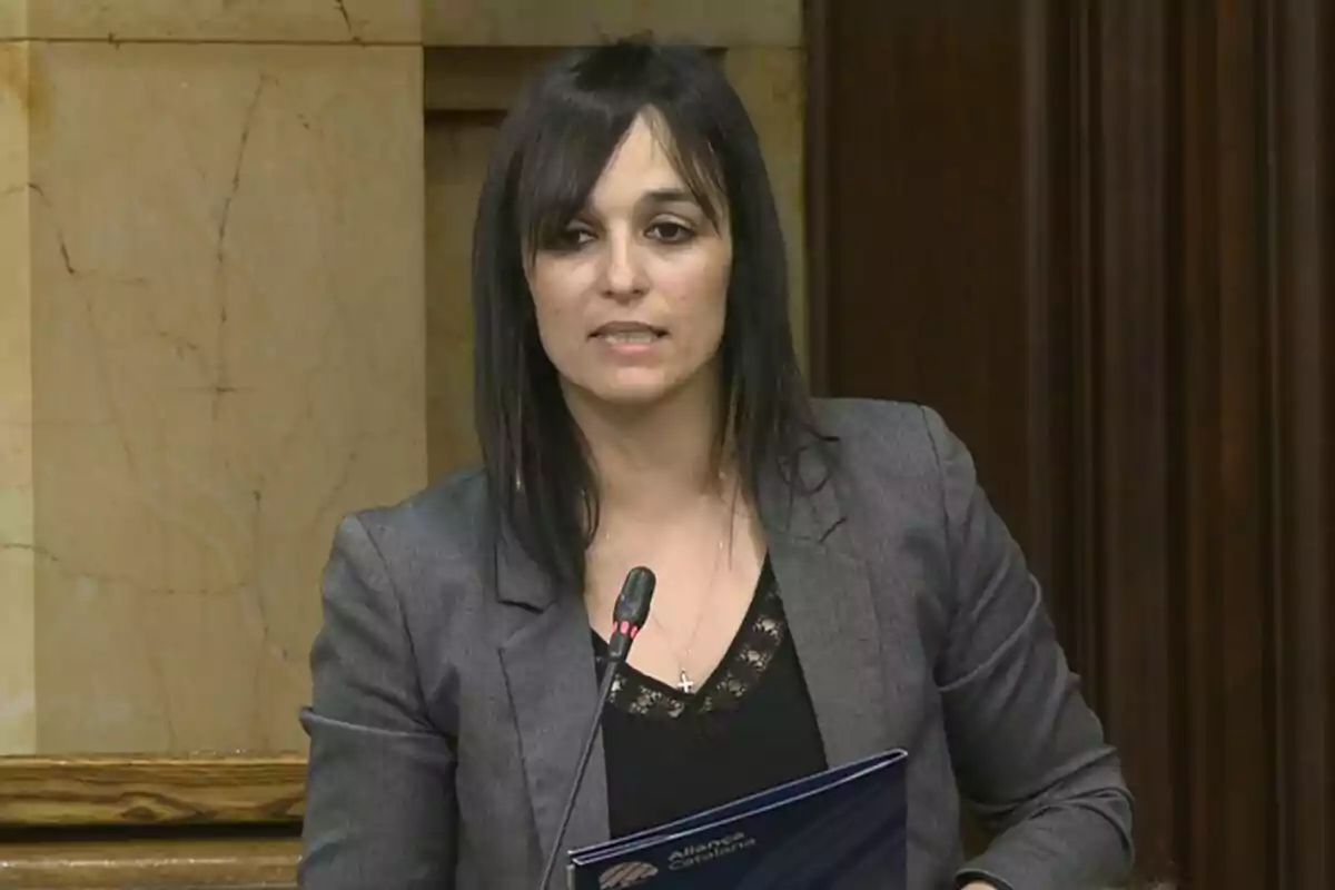 Mujer de cabello oscuro y liso hablando frente a un micrófono en un entorno institucional con paredes de mármol y madera, vestida con saco gris y blusa negra, sosteniendo una carpeta azul