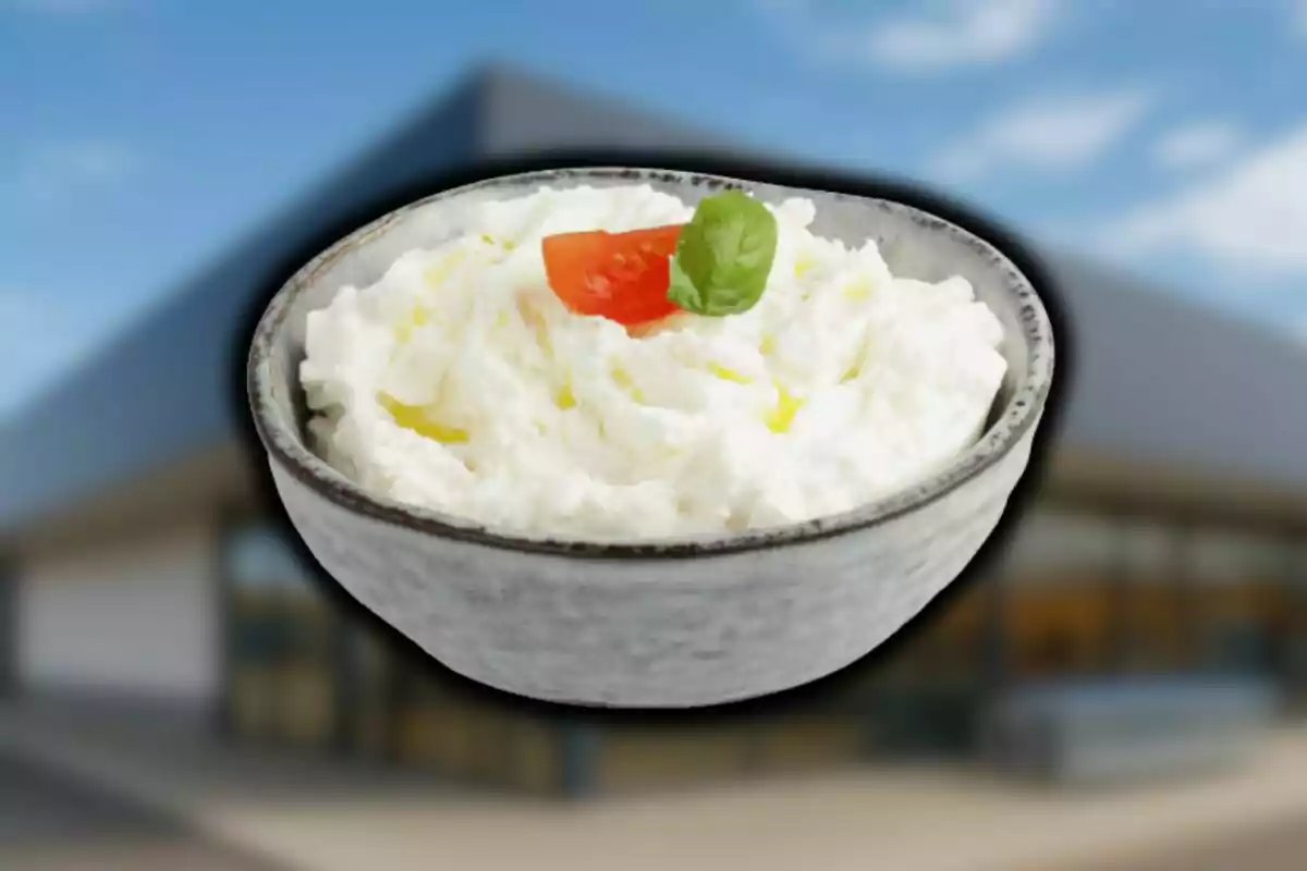 Un bol de formatge burrata d’Aldi decorat amb un tros de tomàquet i una fulla d’alfàbrega sobre un fons desenfocat d’un edifici modern.