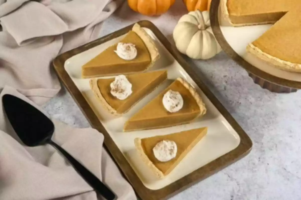 Four slices of pumpkin pie with whipped cream on a rectangular tray next to a black spatula and decorative pumpkins.