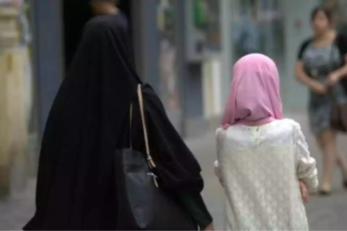 Dos personas caminando por la calle, una vestida con un velo negro y la otra con un velo rosa y ropa clara