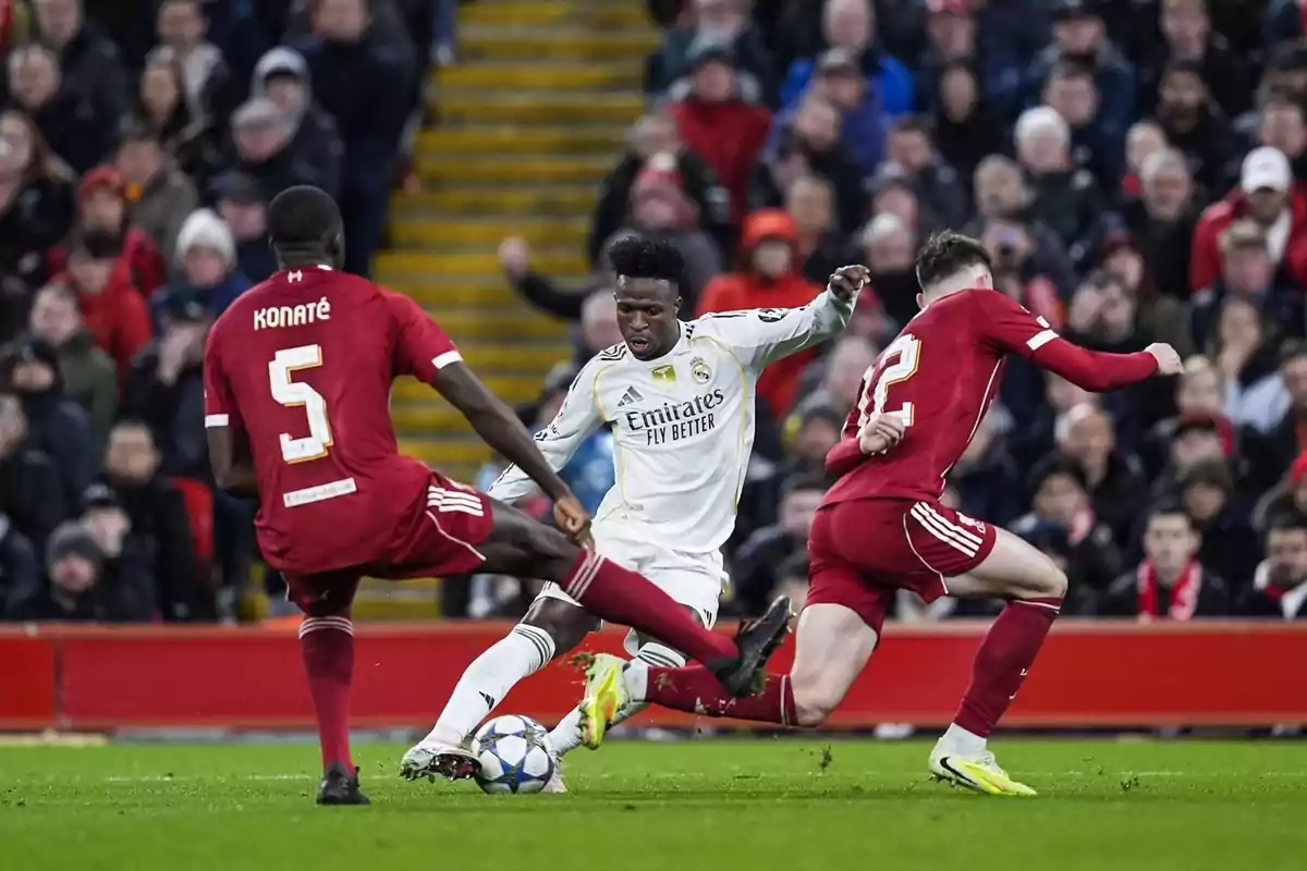 Un jugador del Reial Madrid intenta avançar amb la pilota mentre dos defensors del Liverpool intenten aturar-lo durant un partit de futbol amb el públic observant des de les grades.