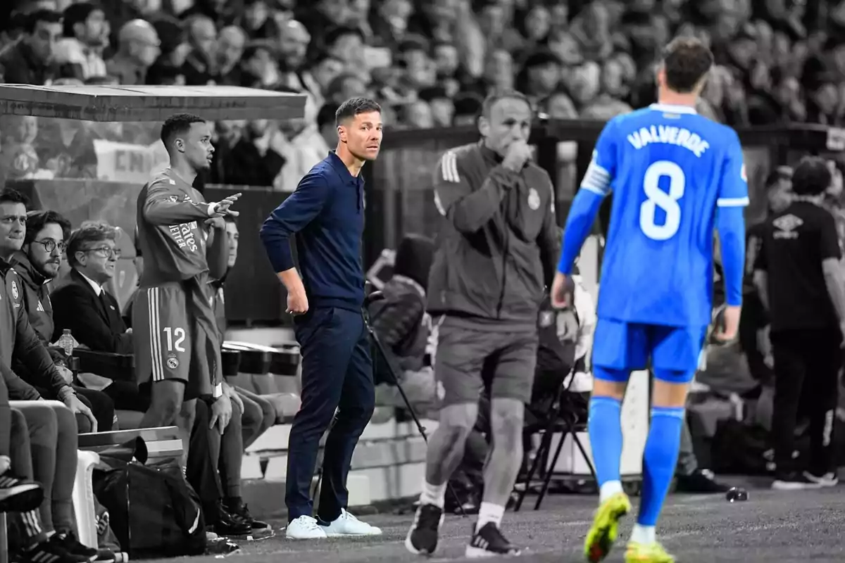 Un entrenador de fútbol observa el partido desde la línea de banda mientras un jugador con el uniforme azul y el número 8 se acerca, el fondo y otras personas están en blanco y negro destacando solo a los protagonistas en color. Un entrenador de fútbol observa el partido desde la línea de banda mientras un jugador con el uniforme azul y el número 8 se acerca, el fondo y otras personas están en blanco y negro destacando solo a los protagonistas en color.