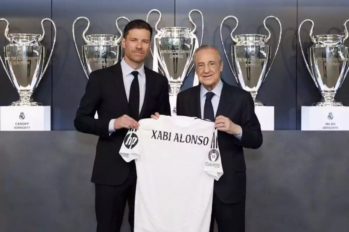 Dos homes amb vestit sostenen una samarreta blanca amb el nom de Xabi Alonso davant de diversos grans trofeus de futbol.