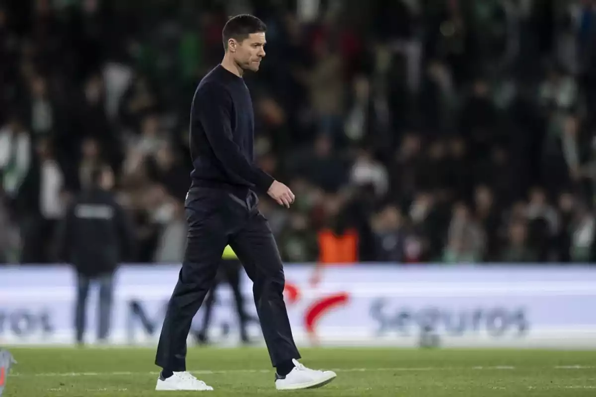 Hombre caminando pensativo sobre el césped de un estadio de fútbol con público desenfocado al fondo
