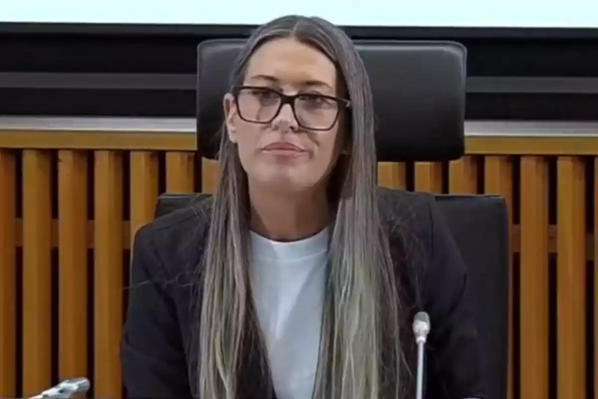 Dona de cabell llarg i llis amb ulleres grosses asseguda davant d’un micròfon en una sala de reunions