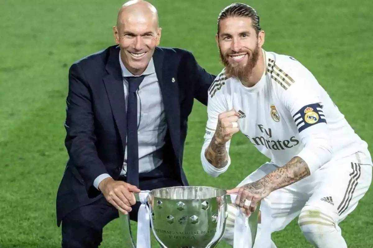Two people smile and pose on a soccer field next to a silver trophy.