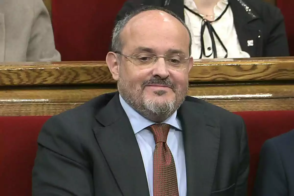 Home de mitjana edat amb barba i ulleres assegut en un banc de fusta amb respatller vermell, vestit amb un vestit fosc, camisa blava i corbata vermella