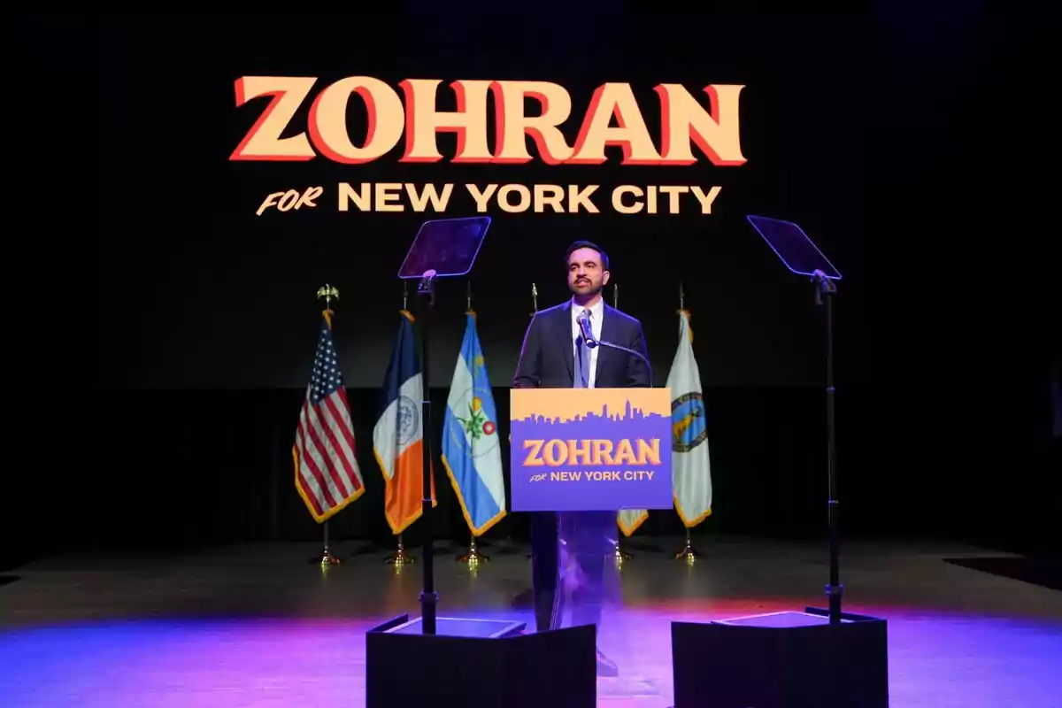 Zohran Mamdani está de pie en un escenario detrás de un podio con el nombre Zohran y el texto for New York City, al fondo hay varias banderas y una pantalla grande con el mismo nombre y mensaje. Zohran Mamdani está de pie en un escenario detrás de un podio con el nombre Zohran y el texto for New York City, al fondo hay varias banderas y una pantalla grande con el mismo nombre y mensaje.