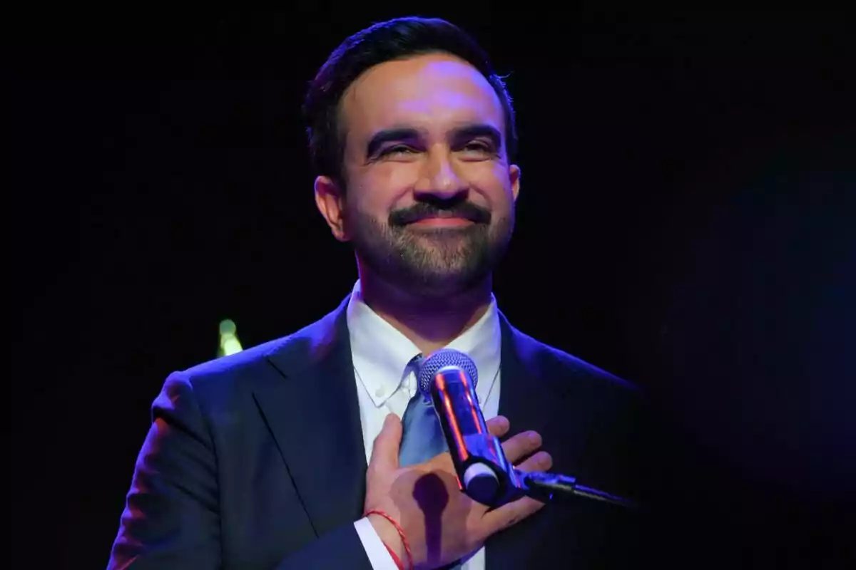 Zohran Mamdani sonríe mientras sostiene una mano en el pecho y está frente a un micrófono, vestido con traje y corbata, con fondo oscuro. Zohran Mamdani sonríe mientras sostiene una mano en el pecho y está frente a un micrófono, vestido con traje y corbata, con fondo oscuro.