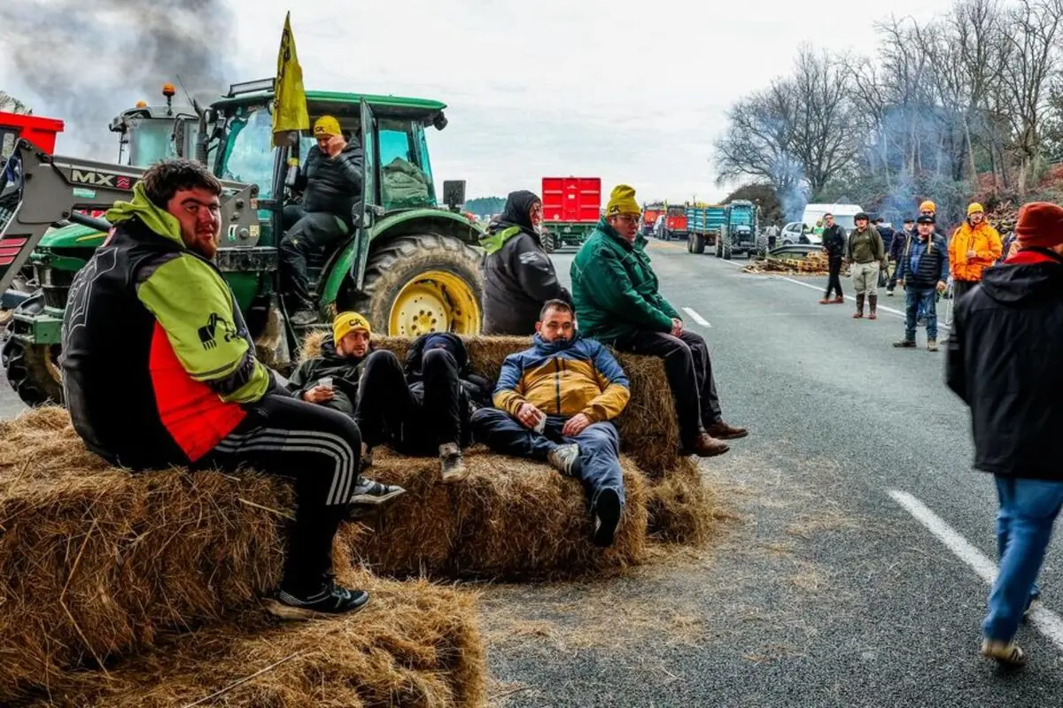 Grupo de agricultores descansando sobre pacas de heno mientras bloquean una carretera con tractores y remolques durante una protesta rural