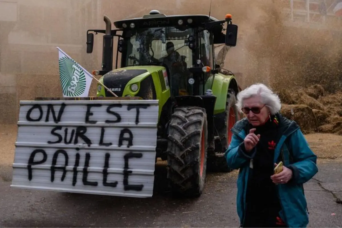 Tractor verde en una protesta agrícola con un gran cartel en francés que dice ON EST SUR LA PAILLE y una mujer mayor caminando delante