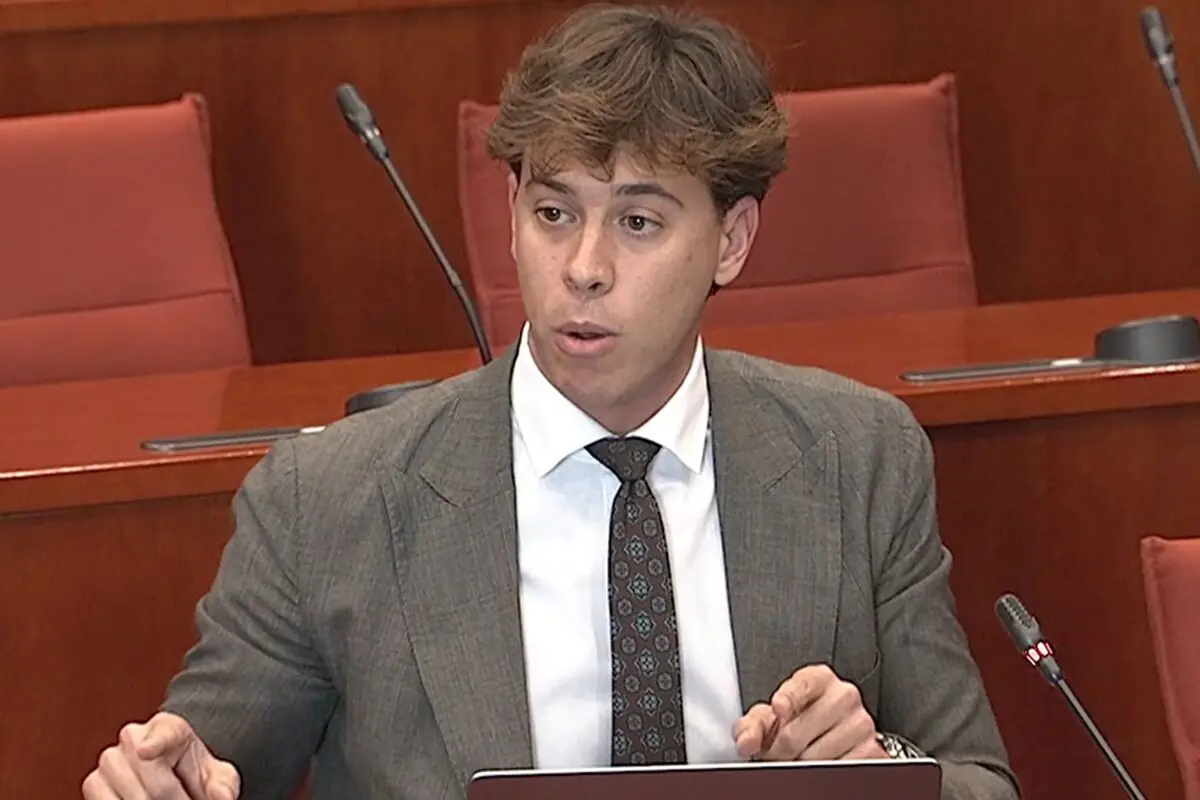 Hombre joven con traje gris y corbata hablando en una sala de conferencias con micrófonos y asientos rojos de fondo