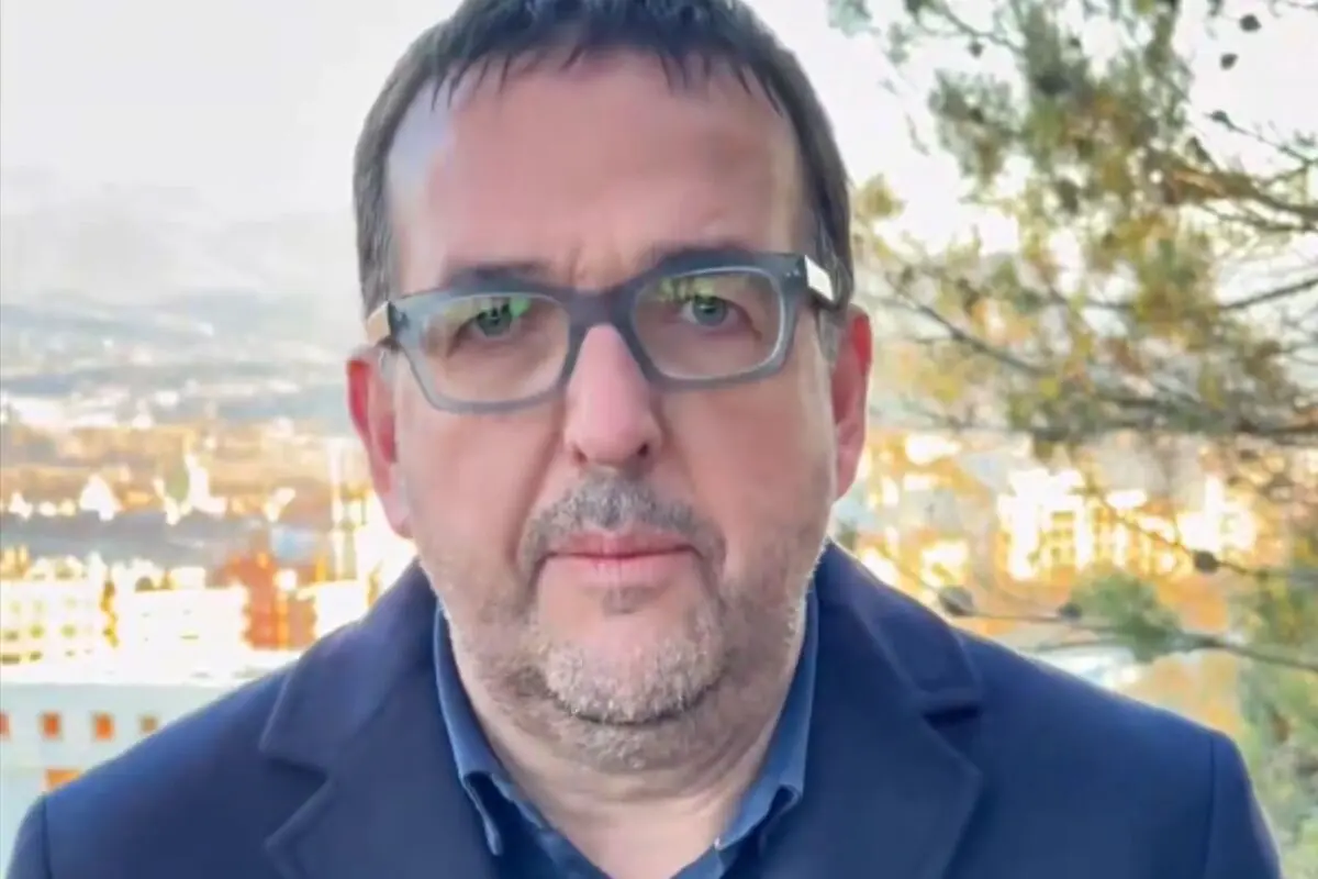 Hombre de mediana edad con gafas y barba corta, vestido con chaqueta azul, al aire libre con un fondo urbano y árboles desenfocados.