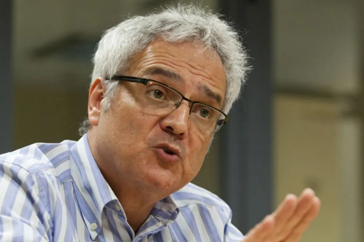 Hombre de cabello canoso y gafas hablando y gesticulando con la mano mientras viste una camisa de rayas azules