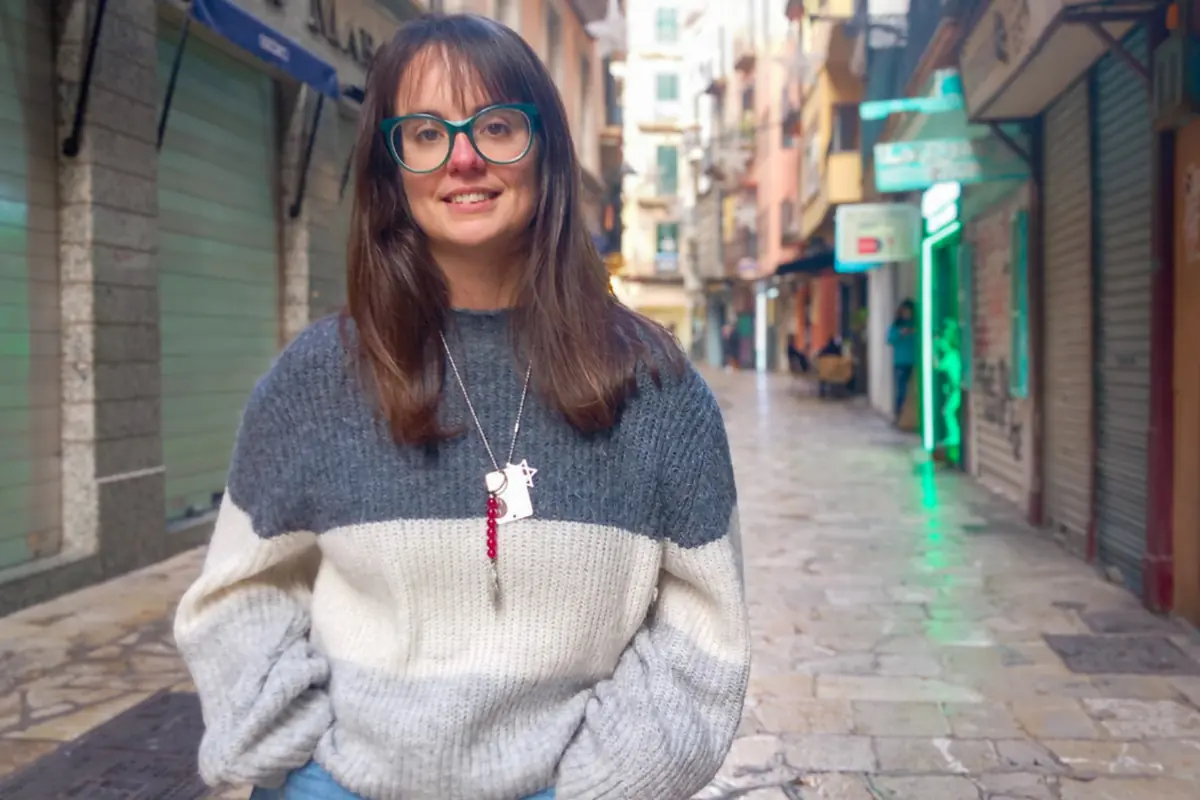 Mujer joven con gafas grandes y suéter gris posando en una calle peatonal con locales cerrados y luces de neón al fondo