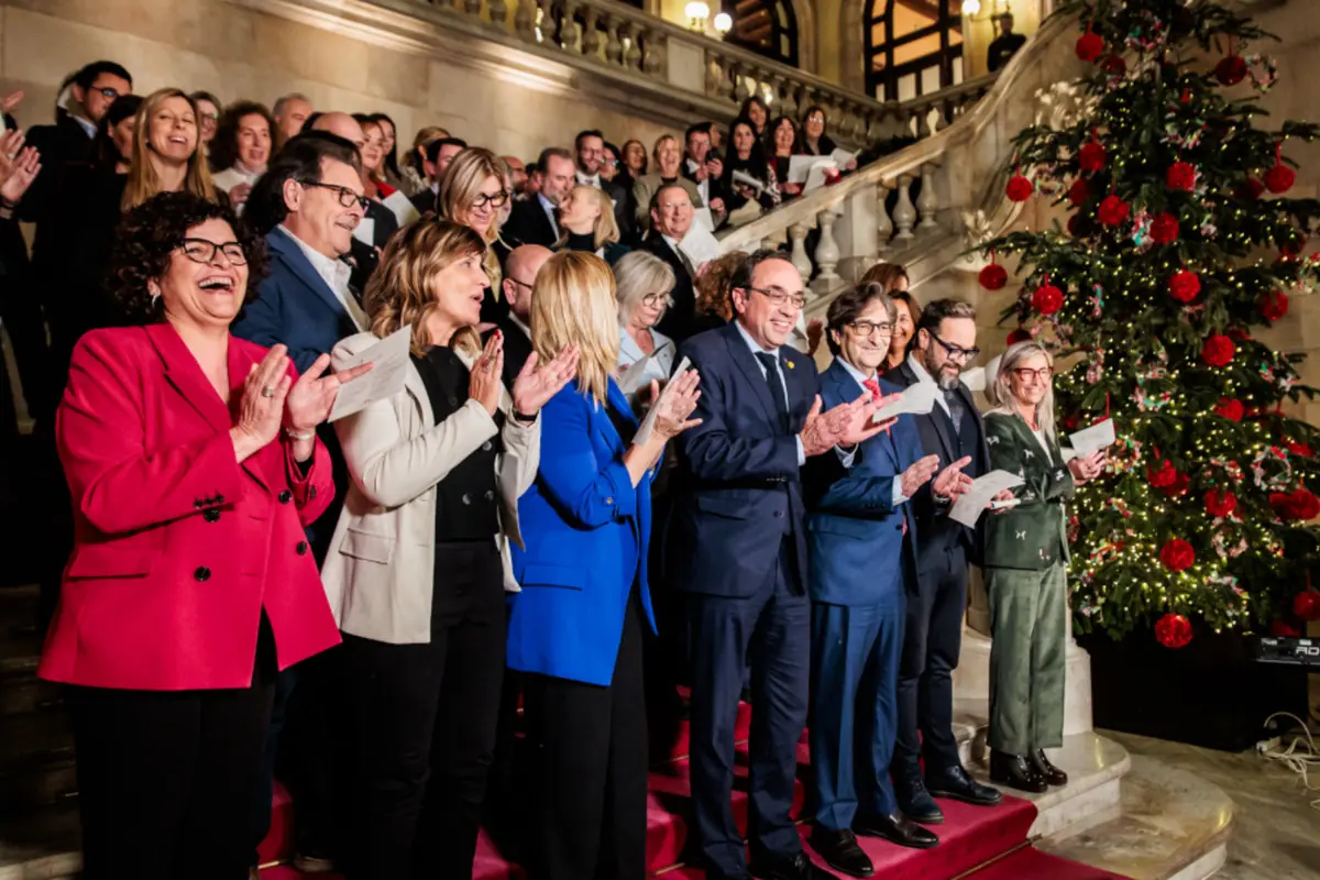 Grupo de personas elegantemente vestidas aplaudiendo y cantando en una escalera decorada con un gran árbol de Navidad iluminado al fondo