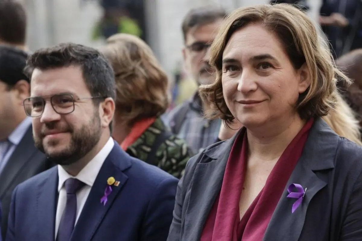 Dos personas vestidas de forma formal en un acto público al aire libre con lazos morados en la solapa rodeadas de más asistentes en segundo plano