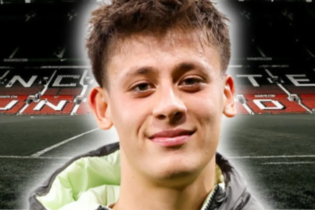 Young man in a sports jacket smiling in front of a soccer stadium with red stands and white letters in the background