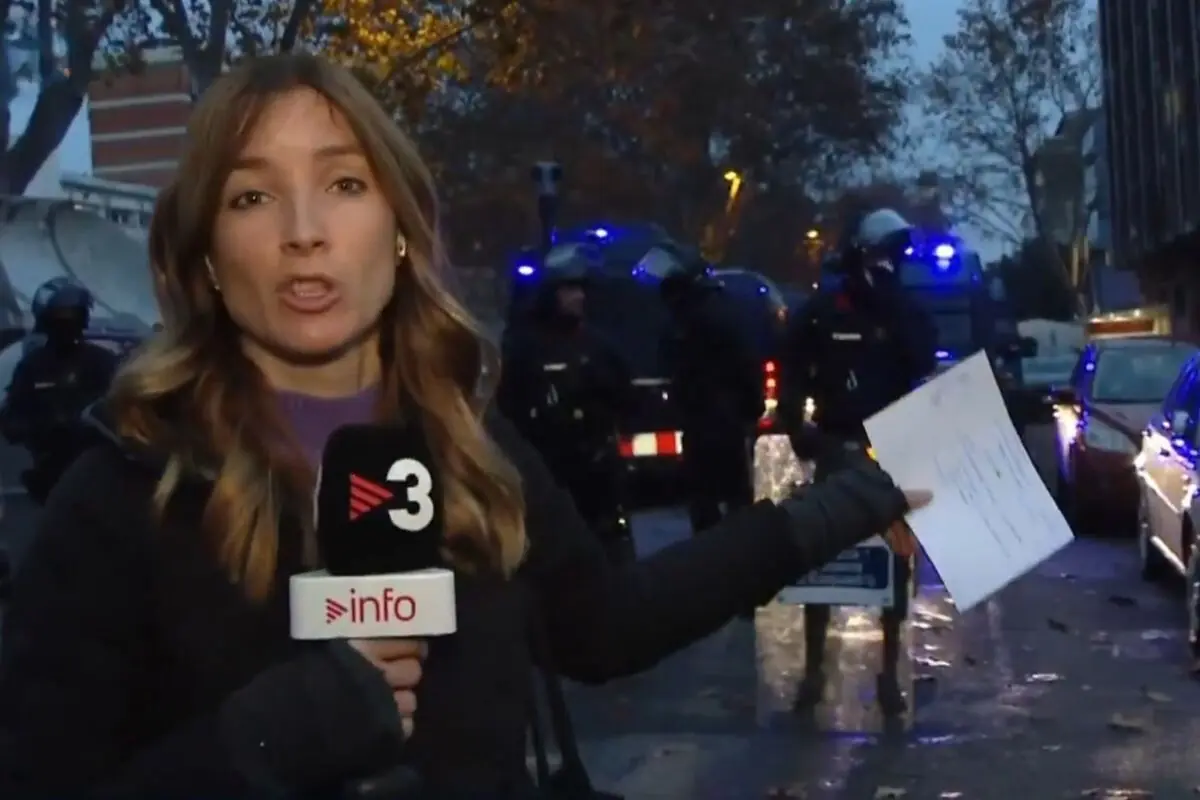 Reportera de televisión informando en la calle con un micrófono de TV3 mientras señala un documento frente a un operativo policial con furgones y agentes al fondo