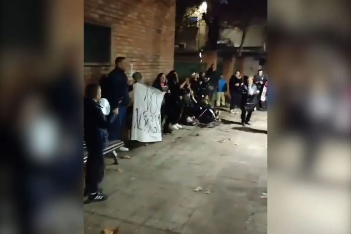 Grupo de vecinos protestando de noche en una calle junto a una pared de ladrillo mientras sostienen una pancarta blanca con un mensaje escrito a mano