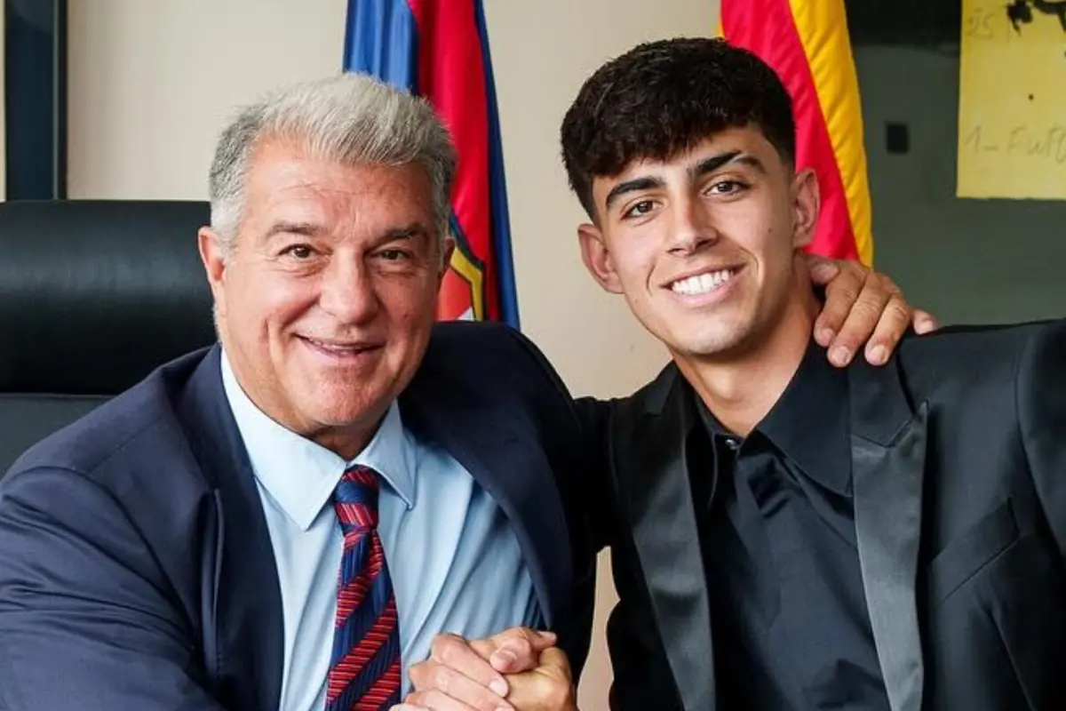 Dos hombres sonríen y se dan la mano mientras posan juntos, ambos vestidos de traje, con banderas de fondo.