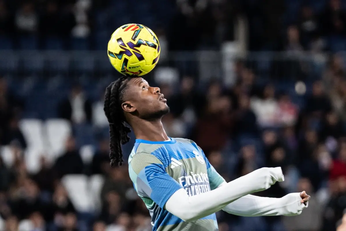 Futbolista con camiseta de entrenamiento azul controlando un balón amarillo con la cabeza en un estadio lleno de público