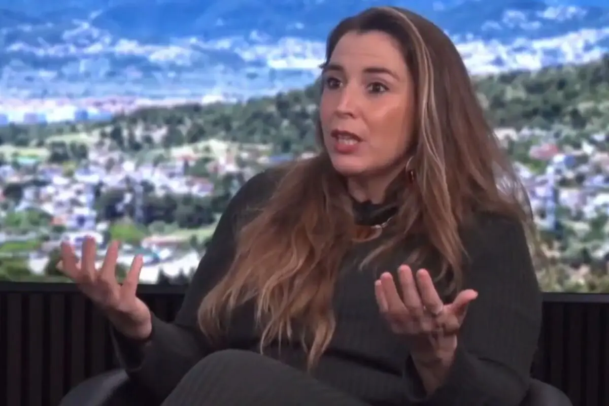 Mujer de cabello largo castaño sentada en un estudio de televisión gesticulando con ambas manos mientras habla con un paisaje urbano desenfocado de fondo
