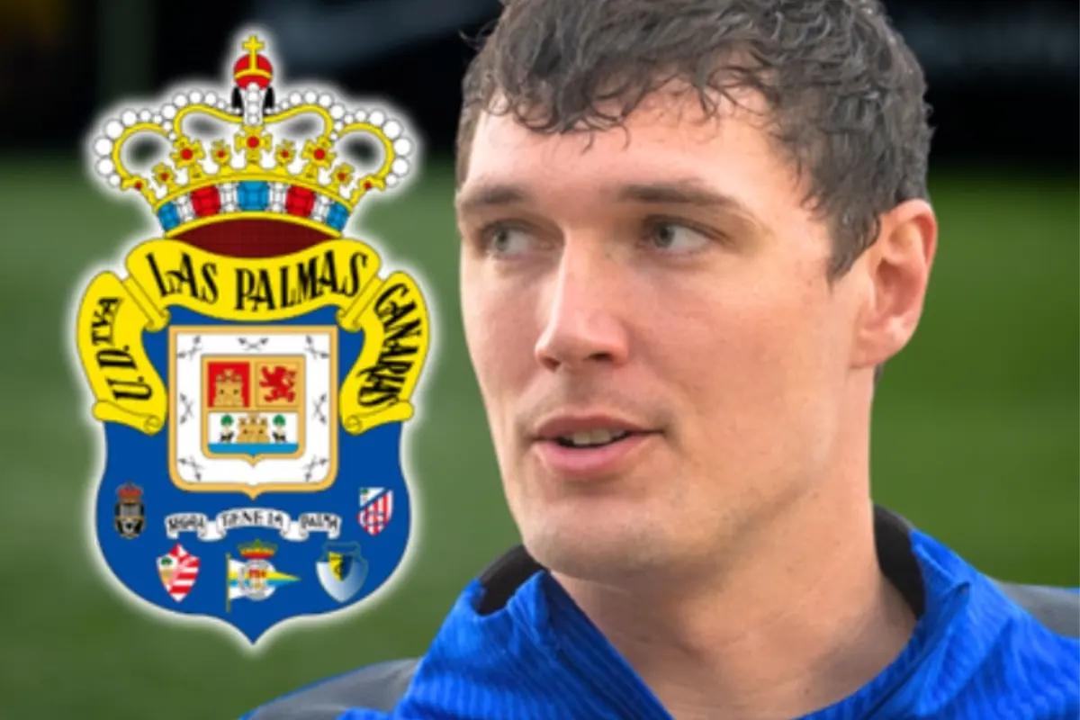 Futbolista amb samarreta blava en primer pla al costat de l’escut del club UD Las Palmas sobre un fons de camp verd