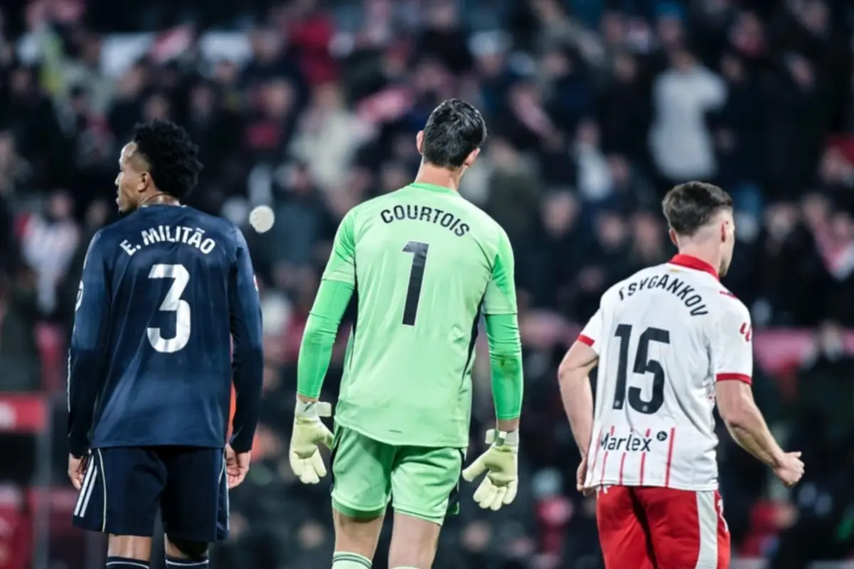 Tres futbolistes d’esquena al camp durant un partit, un amb uniforme blau fosc, un altre amb uniforme verd de porter i el tercer amb uniforme blanc i vermell, amb públic desenfocat al fons