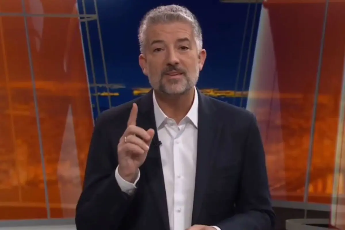 Hombre de cabello canoso y barba corta usando saco oscuro y camisa blanca, levantando el dedo índice mientras habla en un estudio de televisión con fondo naranja y azul