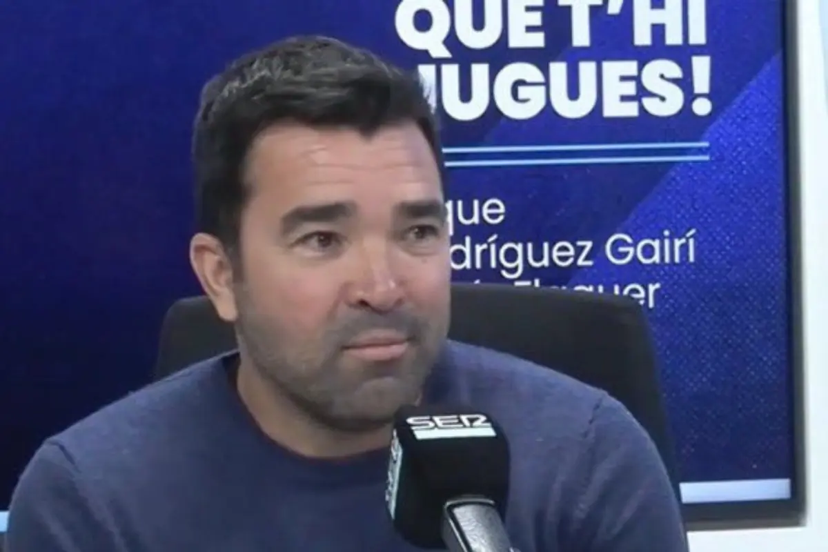 Home de cabells foscos i barba curta parlant davant d’un micròfon en un estudi de ràdio amb fons blau i text a la paret