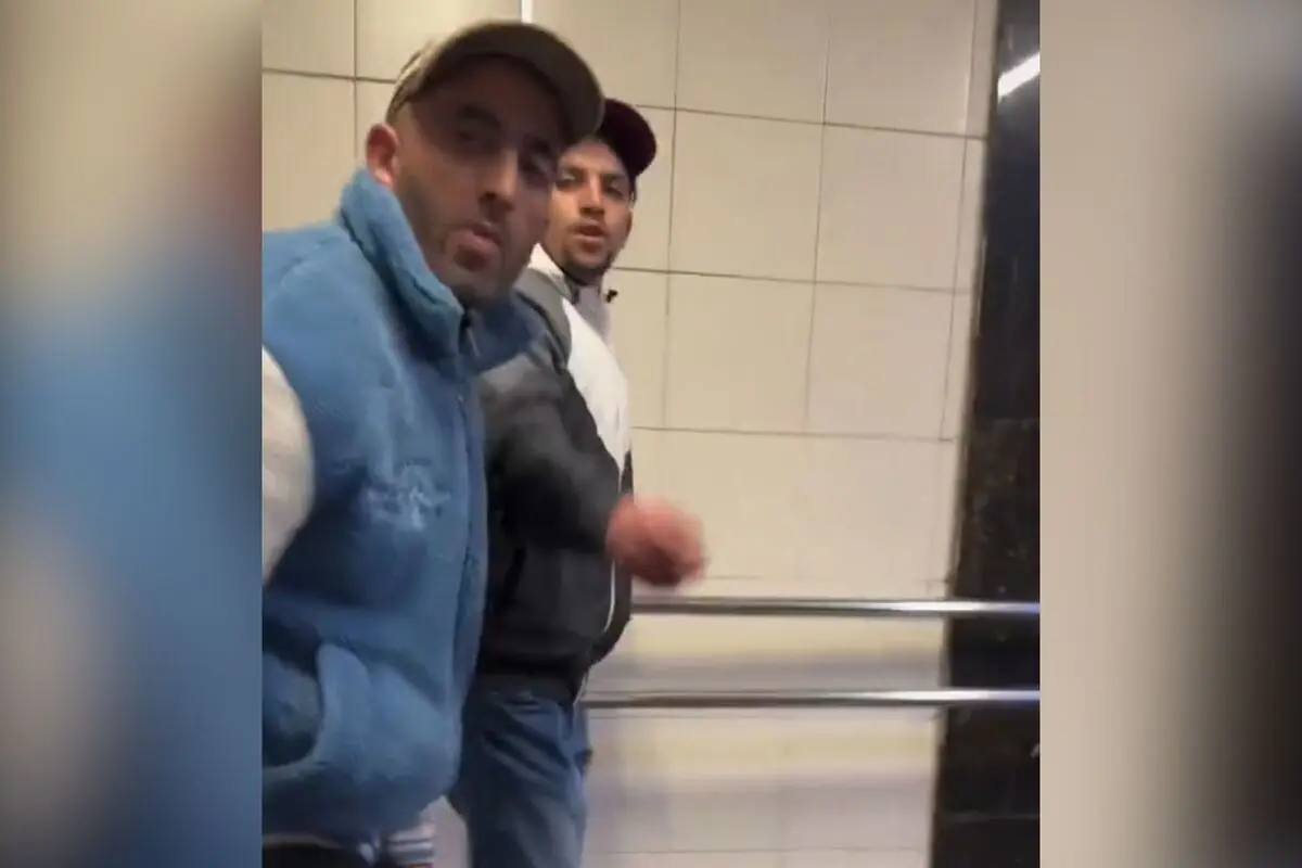 Dos hombres con chaquetas y gorras caminan por un pasillo con paredes de azulejos claros y barandales metálicos.