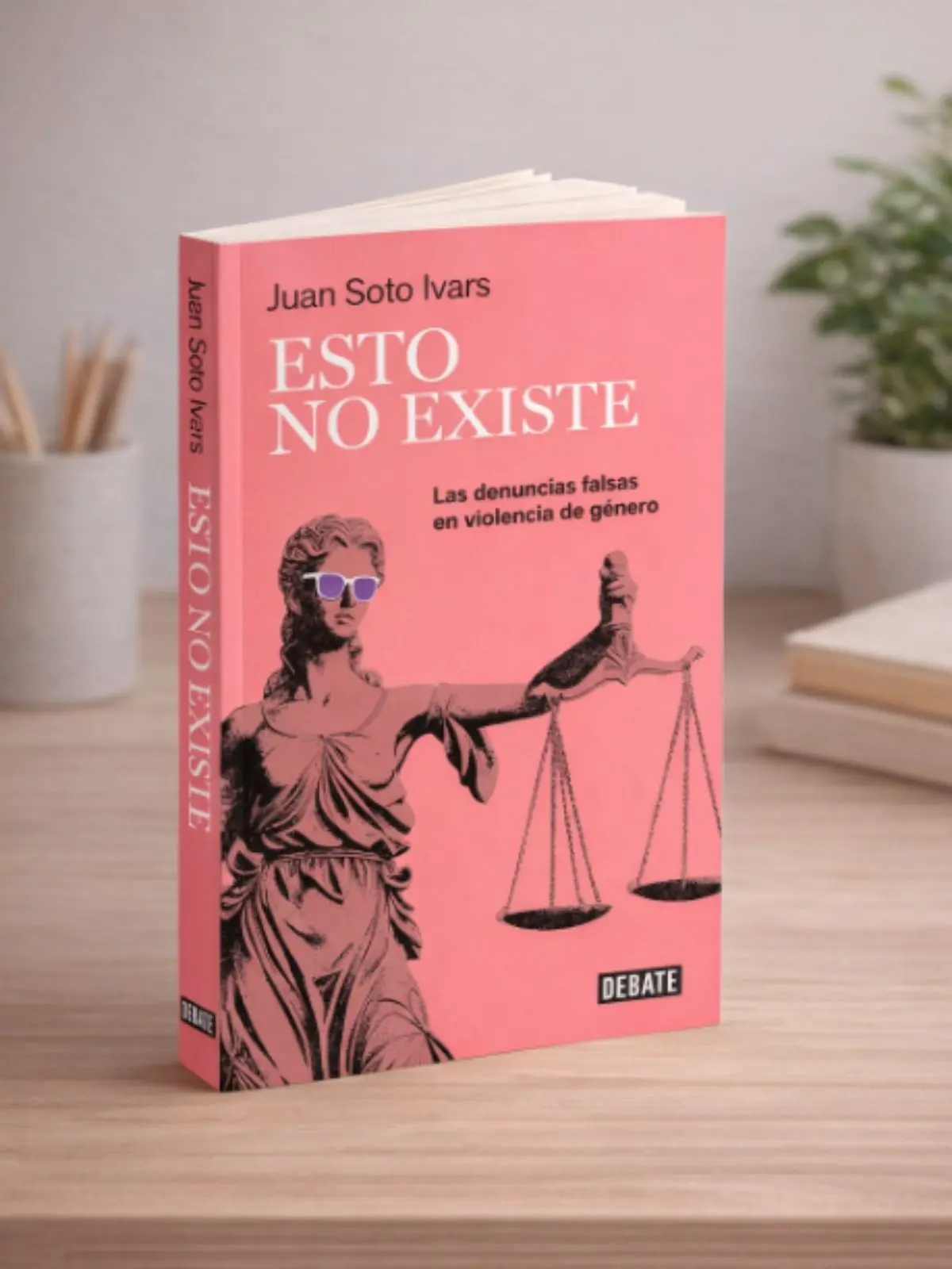 Llibre de tapa tova de color rosa titulat 'Això no existeix' de Juan Soto Ivars recolzat sobre una taula de fusta amb una il·lustració de la deessa de la justícia amb ulleres de sol i balança a la portada.
