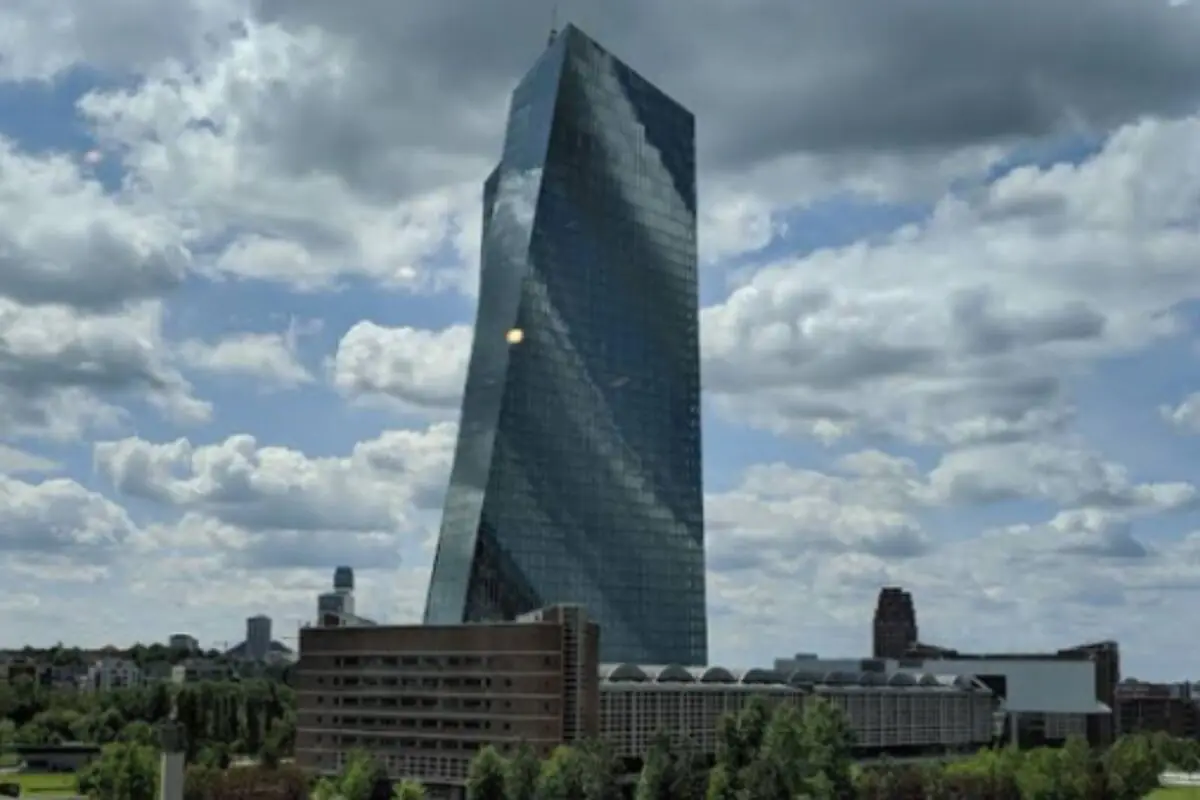 Rascacel de vidre modern amb una forma lleugerament torçada sobre un complex d’edificis baixos i arbres sota un cel ennuvolat; es tracta de l’edifici del BCE.