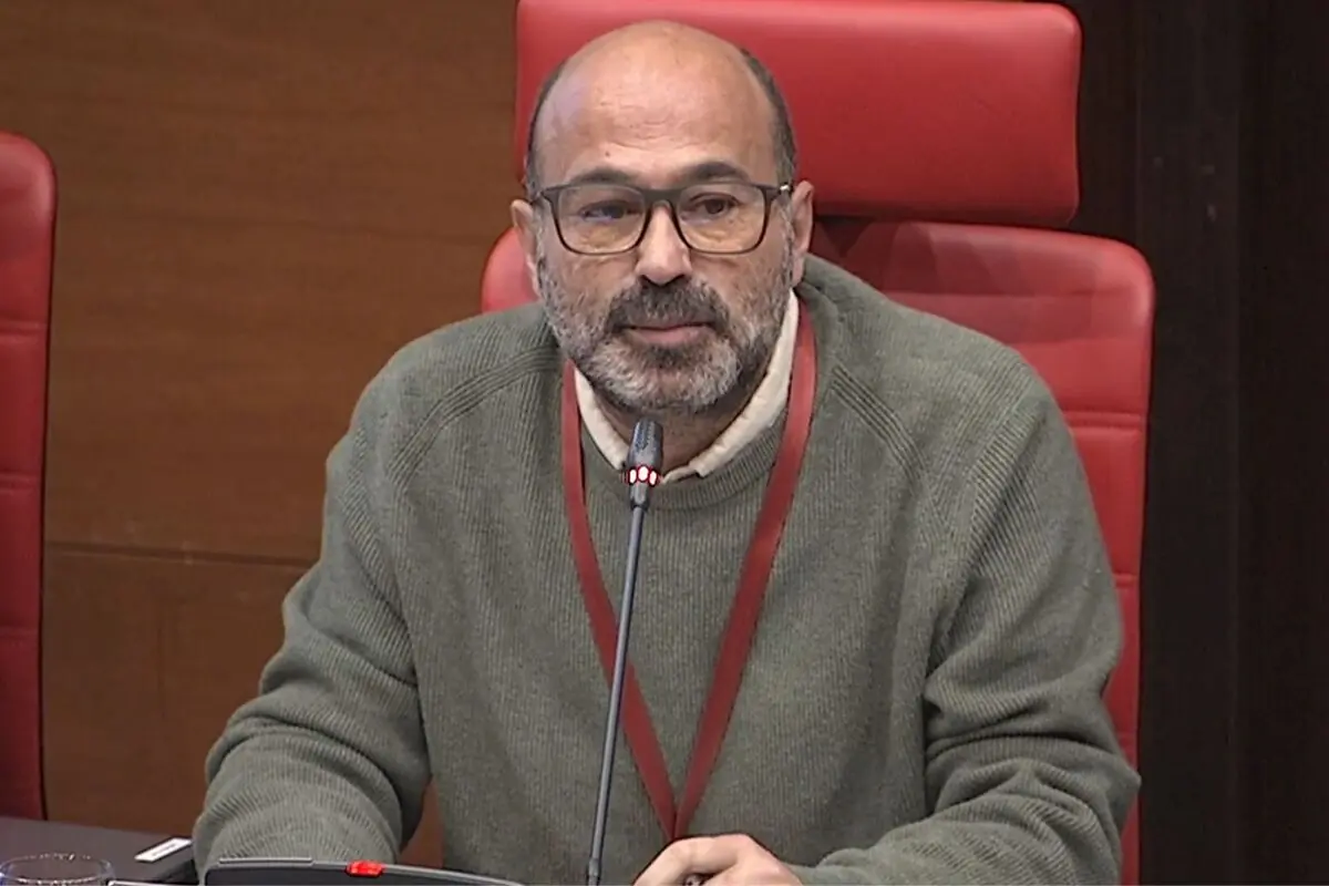 Hombre calvo con gafas y jersey verde hablando frente a un micrófono en una sala de conferencias con asiento rojo