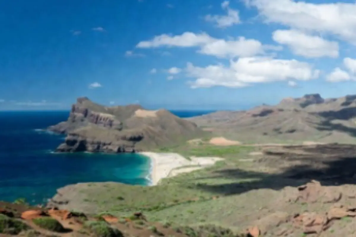 Vista panoràmica d’una platja de sorra blanca envoltada de muntanyes i mar blau sota un cel parcialment ennuvolat a les Illes Canàries.